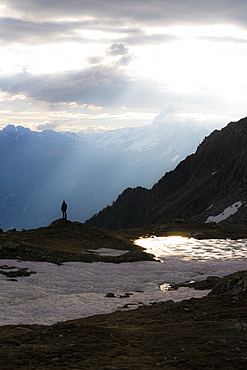 Silhouette of man standing under sun rays at dawn on shores of lake Zana, Valmalenco, Valtellina, Lombardy, Italy, Europe