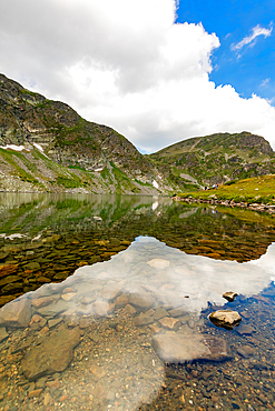 Vertical, eye-level view — Clear mountain lake with submerged rocks visible, Rila Mountains, Bulgaria. Snow patches on surrounding slopes under partly cloudy sky.