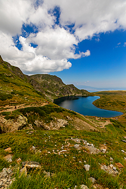Vertical, eye-level view — Heart-shaped Kidney Lake, Rila Mountains, Bulgaria. Foreground slope with rocky terrain; lake below surrounded by grassy hills.