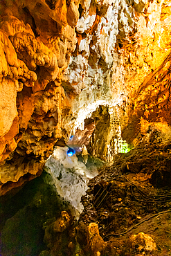 Vertical ground-level shot inside a cave at Matka Canyon, featuring rugged rock formations illuminated by warm artificial lighting with hints of natural green light in the background.