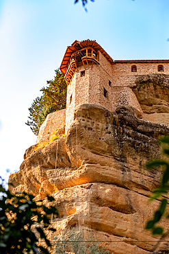 Vertical ground-level shot looking up at a stone monastery tower built on the edge of a steep rock cliff, with warm sunlight highlighting the textures and a soft blue sky above.