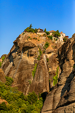 Vertical ground-level shot showing a monastery perched atop a steep rock face, with rugged cliff textures and sparse greenery under a clear blue sky. The framing emphasizes the vertical rise of the formation.