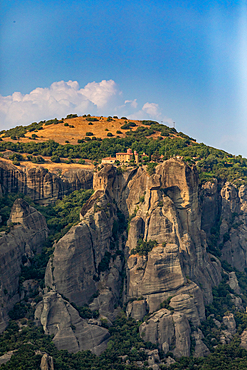 Vertical orientation view of a Meteora monastery built on a high rocky cliff, with a grassy hilltop and scattered trees in the background, Thessaly, Greece.