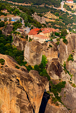 Aerial view in vertical orientation of a Meteora monastery built on a steep rock formation, surrounded by cliffs, greenery, and distant buildings, Thessaly, Greece.