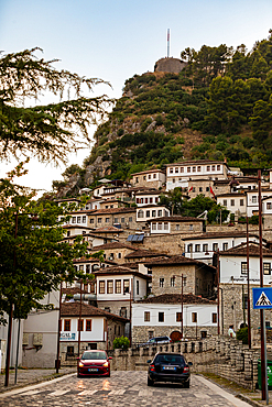 Cars drive through the cobbled streets of Berat, Berat County, Albania, beneath rows of traditional Ottoman-style houses leading up to the historic hilltop Kala Castle.