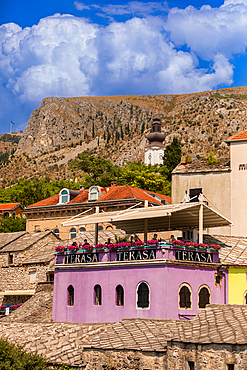 A vibrant purple rooftop café in Mostar’s Old Town contrasts with traditional stone buildings, while an Orthodox church tower rises in the distance against the rocky hillside.