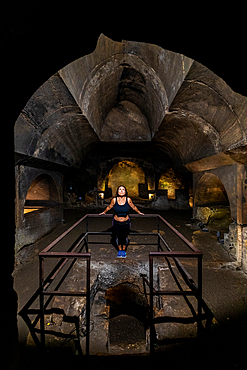 A woman stands at the center of the medieval underground Catacombs of Jajce, a carved stone crypt and chapel illuminated by soft lighting within the historic rock-hewn complex.