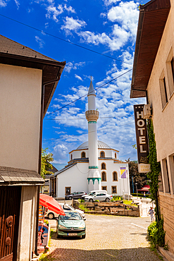 A street-level view of a white mosque with green accents in Jajce, Federation of Bosnia and Herzegovina, Bosnia and Herzegovina. The narrow street scene includes traditional buildings, a hotel sign, and parked cars under a bright summer sky.