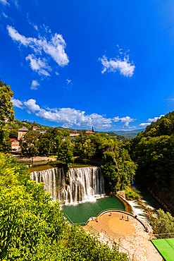 Aerial view of Pliva Waterfall and circular viewing platform in Jajce, Federation of Bosnia and Herzegovina, Bosnia and Herzegovina. The urban waterfall sits below the historic town and is surrounded by lush greenery and summer skies.