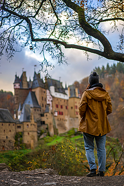 Back view of young woman with yellow jacket looking at Eltz medieval historic castle in an autumn landscape with trees at sunrise, Wierschem, Rhineland-Palatinate, Germany, Europe