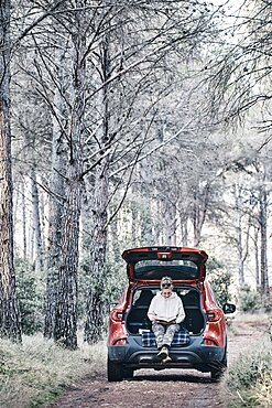 Young adult caucasian woman reading a book on the open trunk of a car parked in a forest path. Travels concept. Ayegui, Navarre, Spain, Europe.