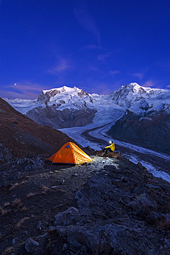 Hiker with head lamp sitting beside the tent in front of Monte Rosa massif, Gorner glacier (Gornergletscher) and Lyskamm peaks at dusk, Riffelsee, Zermatt, canton of Valais, Swiss Alps, Switzerland, Europe