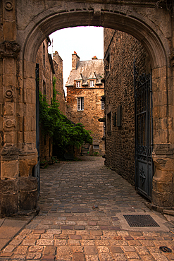 A historic stone archway opens onto a narrow cobbled street with medieval houses in the old town of Dinan, Brittany, France.