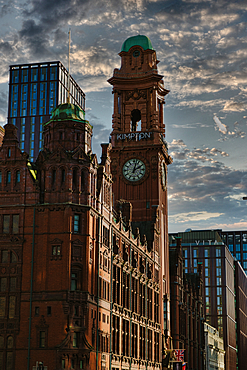 A tall, brick building with a clock tower and green roof, glass-fronted skyscraper under a cloudy sky in Manchester, UK.