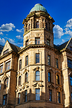 A tall, ornate building with a domed roof and numerous windows. The building is made of light-colored stone and has a symmetrical design. The sky is blue with white clouds, and there are shadows cast by the building on the facade in Manchester, UK.