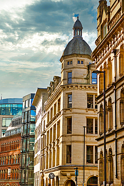 A city streetscape featuring several multi-story buildings of varying architectural styles, including a prominent building with a cupola, under a partly cloudy sky. The buildings are primarily light beige and tan, with one modern glass building.