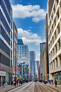 A city street scene featuring modern high-rise buildings, pedestrians, and tram tracks. The architecture is primarily glass and concrete, with a mix of styles. The sky is partly cloudy, adding depth to the image in Manchester, UK.