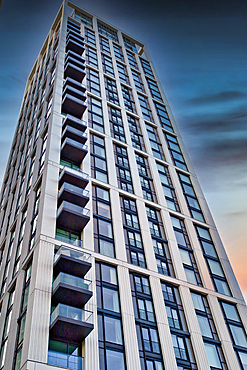 A tall, modern residential building with numerous balconies and large windows is captured against a twilight sky. The building's light-colored facade contrasts with the dark window frames and balconies in London, UK.