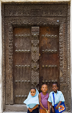 Three children sitting in front of a carved wooden Arab door in Stone Town, Zanzibar, Tanzania, East Africa