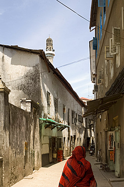 A woman in a colourful headscarf walking down a narrow street with a mosque minaret in Stone Town, Zanzibar, Tanzania, East Africa, Africa