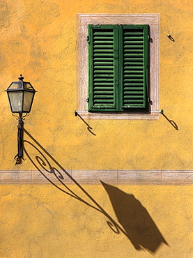 Architectural detail showing green wooden shutter on yellow wall with ornate wall lamp and shadow, San Quirico d'Orcia, Tuscany, Italy, Europe