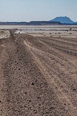 Four by four vehicle in the distance on long straight gravel road, blue mountains in the background, north of Solitaire, Namibia, Africa