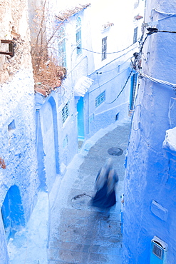 Woman in dark blue traditional clothing running down (blurred) a typical blue painted alleyway in Chefchaouen, Morocco, North Africa, Africa