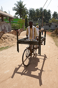 Man cycling through village street with two metal bed frames balanced on his bicycle, Hirapur, Orissa, India, Asia