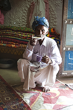 Turbanned man sipping tea from saucer in tribal home in front of piles of hand embroidered quilts, Soyla, Kachchh, Gujarat, India, Asia