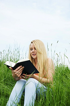 Young blonde woman sitting on a lawn, reading a book