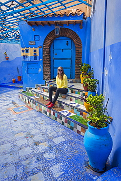 Young woman in the old town sits on a staircase, blue house walls, medina of Chefchaouen, Chaouen, Tangier-Tétouan, Kingdom of Morocco