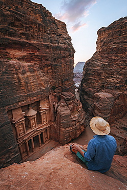 Tourist sits at the edge of a rock and looks from above into the canyon Siq, Pharaoh's treasure house beaten into the rock, facade of the treasure house Al-Khazneh, Khazne Faraun, Mausoleum, Nabataean city Petra, near Wadi Musa, Jordan, Asia