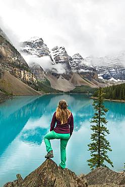 Young woman standing in front of a lake looking into mountain scenery, clouds hanging between mountain peaks, reflection in turquoise lake, Moraine Lake, Valley of the Ten Peaks, Rocky Mountains, Banff National Park, Province of Alberta, Canada, North America