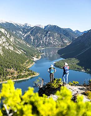 Two hikers look into the distance over the Lake Plansee, surrounded by mountains, Schoenjoechl, Plansee, Tyrol, Austria at the back.