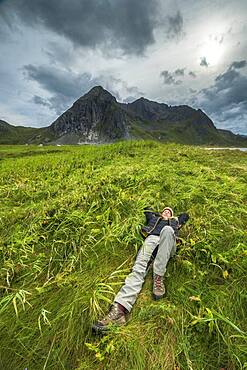 Woman lies pleasurably in a meadow in front of threatening mountain range, Skagsanden, Flakstad, Lofoten, Nordland, Norway, Europe