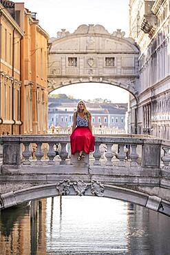 Young woman with red skirt, tourist sitting on a bridge railing, bridge over the Rio di Palazzo, behind Bridge of Sighs, Venice, Veneto, Italy, Europe