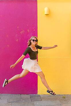 Young woman in dress jumps happily in front of colorful house, yellow and pink house facade, Burano Island, Venice, Veneto, Italy, Europe