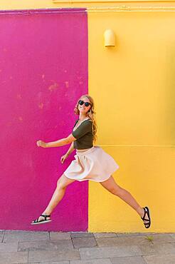 Young woman in dress jumps happily in front of colorful house, yellow and pink house facade, Burano Island, Venice, Veneto, Italy, Europe