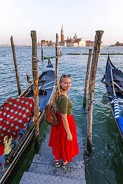 Young woman with red dress on a jetty, Venetian gondolas, in the back church San Giorgio Maggiore, Venice, Veneto, Italy, Europe