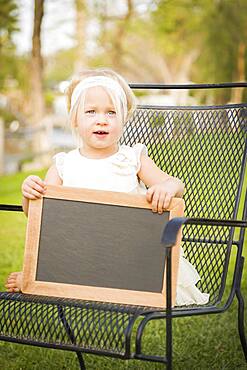 Cute baby girl sitting in chair holding blank blackboard