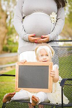 Pregnant mom behind cute baby girl sitting in chair holding blank blackboard