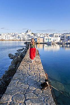 Young woman with red dress on harbour wall, behind church of Naoussa, harbour town Naoussa, island Paros, Cyclades, Greece, Europe