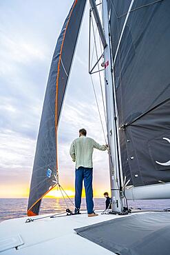 Young man standing leaning against the mast on a catamaran, looking into the sunset, Rhodes, Dodecanese, Greece, Europe