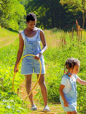 Short haired young african woman and daughter watering vegetable garden with hose taking care of vegetables in the organic farm wearing denim dungarees