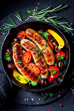 Grilled sausages and vegetables with herbs in iron pan on black background