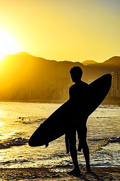 Surfer silhouette with his longboard looking at the Iapnema beach waves in Rio de Janeiro during sunset, Brasil