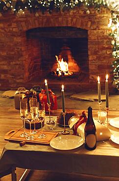 Christmas or New year dinner against burning fireplace and xmas gifts in cozy house decorated with led lights, festive table with freshly baked cake, lit candles, glasses of champagne, selective focus