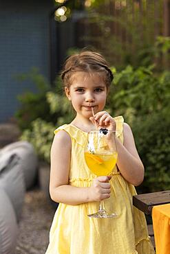 Portrait of little girl drinking juice in a glass, decorated with fruits, with straw at outdoor park. Child in summer