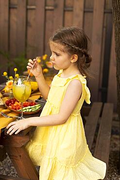 Portrait of little girl drinking juice in a glass, decorated with fruits, with straw at outdoor park. Child in summer