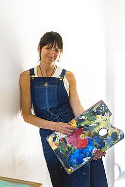 Portrait of a woman artist leaning on the wall, holding a wooden pallete while looking at camera